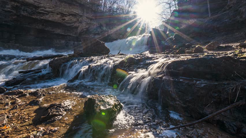Timelapse of waterfall stream flowing down rocks, sun lens flare in background
