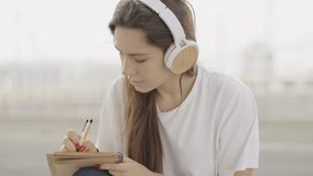 Young woman listening music while writing on notebook outside