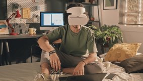 Full medium shot of curly young man in white VR headset turning his head around slowly, sitting on unmade bed, laptop in front of him. Turned-on screen, lamps and window in background, early morning