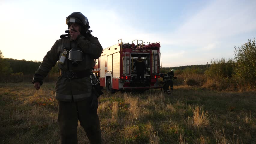 Professional fireman in full equipment talking on walkie-talkie on place at fire near a big red truck. Male firefighter speaking on radio communication against background of fire engine at countryside Royalty-Free Stock Footage #1103859733