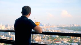 Adult man standing with cocktail at luxury rooftop restaurant with cityscape at evening. Young male in black polo with orange mocktail at sky bar terrace enjoy city skyline and river view. - Powered by Shutterstock - Get 15% off with code: PIKWIZARD15