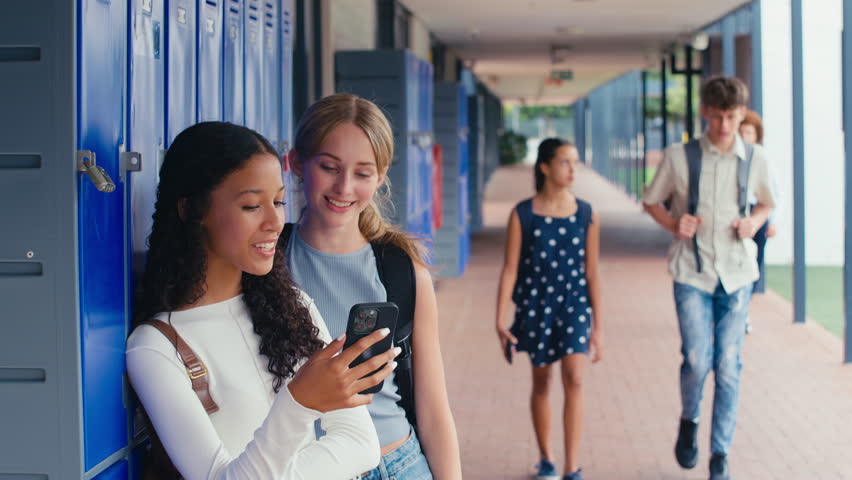 Two female high school or secondary students looking at social media or internet on mobile phone by lockers - shot in slow motion