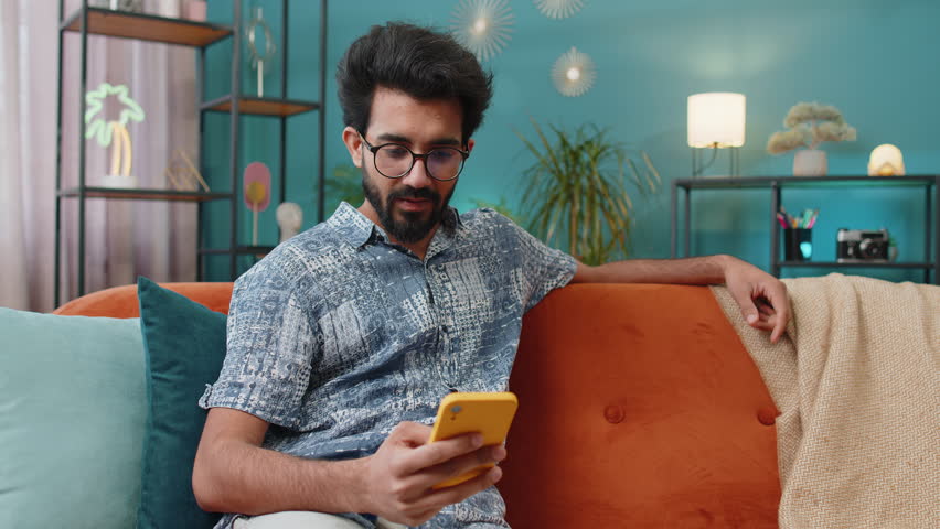 Happy indian man use mobile smartphone typing browsing say Wow yes found out great big win good news celebrate lottery jackpot doing winner gesture. Hindu young guy at home in room sitting on sofa Royalty-Free Stock Footage #1103866873