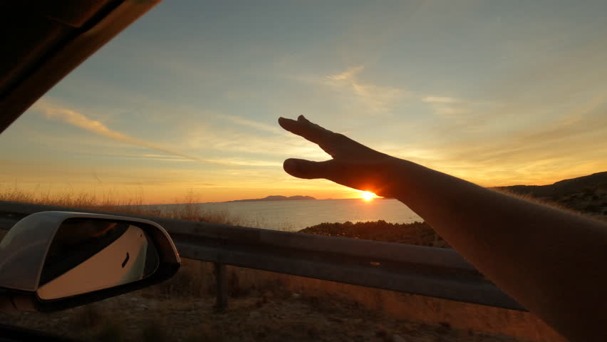 LENS FLARE: Woman's hand waves through window of car driving along the seashore. She enjoys summer breeze and freedom while driving to her summer holiday destination on beautiful coast of Adriatic Sea