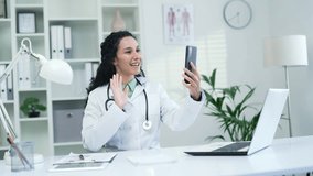 Smiling female doctor in white coat talking on video call using smartphone in modern hospital clinic. A brunette medical worker physician consults a patient sitting at a desk at a workplace in office