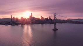 Beautiful aerial view of Bay Bridge with skyscrapers in downtown San Francisco in cinematic rose golden sunset light. Drone flying above water surface along Bay Bridge construction.Scenic cityscape 4K