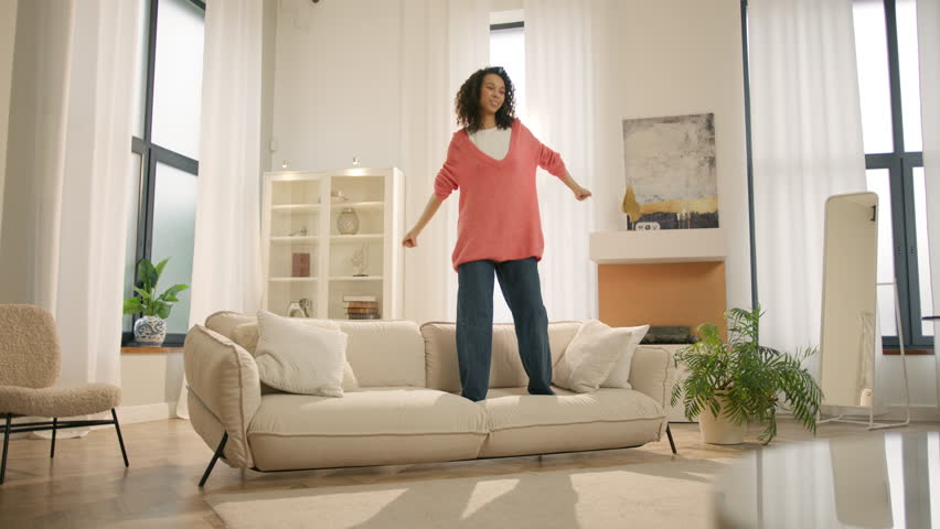 Happy young black woman jumping on sofa, feels overjoyed by celebrating move in day to own home. Mixed race woman laughing enjoying newly rented modern flat or urban apartment. Fashion furniture store Royalty-Free Stock Footage #1103871977