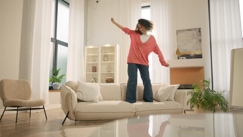 Happy young black woman jumping on sofa, feels overjoyed by celebrating move in day to own home. Mixed race woman laughing enjoying newly rented modern flat or urban apartment. Fashion furniture store | Shutterstock HD Video #1103871977