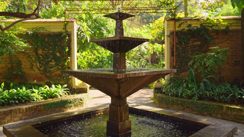 Moving towards an outdoor water fountain feature in slow motion with garden greenery and brick wall in the back 