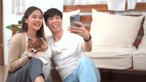 Happy cheerful Asian young couple taking a selfie photography or making a selfie video call on smartphone with their little dog in living room. Man and woman smiling to camera on smartphone.