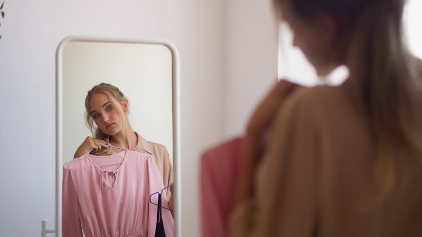 Young woman with ponytail looks at reflection in mirror choosing between elegant pink and classic black dress for evening party slow motion Royalty-Free Stock Footage #1103884051