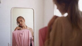 Young woman with ponytail looks at reflection in mirror choosing between elegant pink and classic black dress for evening party slow motion
