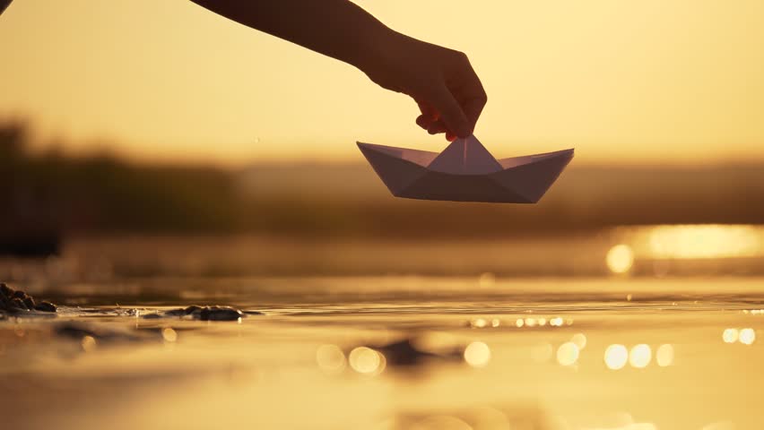 A woman's hand launches white paper craft on water at beach. Paper boat on water. Close-up of girl's hand with boat. Floating paper boat on water. Royalty-Free Stock Footage #1103891923