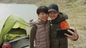 Medium shot of young Asian female friends making selfie or video, one of them holding phone camera, hugging closely, smiling, while hiking on lake coast, their tent in background. Sunny, windy day