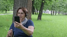 Smiley senior woman having video call while sitting on the grass under the tree in city park.