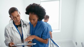 Portrait of smiling female doctor wearing white coat discussing patient notes on clipboard with nurse in scrubs on hospital stairs with male colleagues meeting in background - shot in slow motion - Powered by Shutterstock - Get 15% off with code: PIKWIZARD15