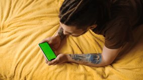 Caucasian man using smartphone in horizontal mode with a green mock-up screen lies on a yellow blanket and watches videos or movies. 4k footage. Focus on Chroma Display and hands.