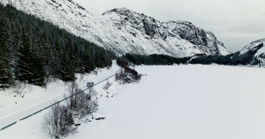 Campervan drives along road through harsh winter snowy terrain, Lofoten. Aerial