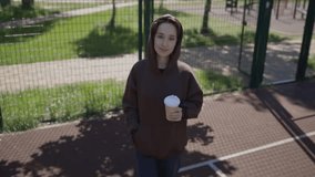Caucasian girl drinks coffee on a sunny day. a girl in a hoodie walks around the sports ground and drinks coffee. Joyful girl in a brown sweatshirt drinks takeaway coffee on the basketball court.