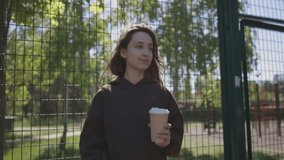 a Caucasian girl drinks coffee in the city. a girl in a hoodie walks along the sports ground and drinks coffee. A girl in a brown hoodie walks through a basketball ground,with a cup of takeaway coffee