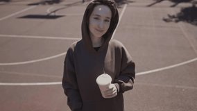 Caucasian girl drinks coffee on a sunny day. a girl in a hoodie walks around the sports ground and drinks coffee. Joyful girl in a brown sweatshirt drinks takeaway coffee on the basketball court.