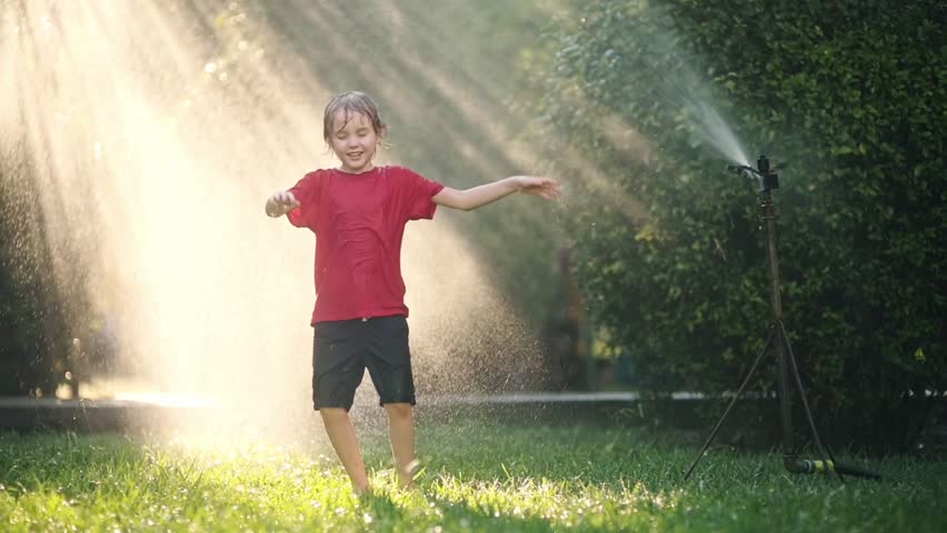Slow motion video of funny little boy playing with garden sprinkler in sunny backyard.Elementary school child laughing, jumping and having fun with spray of water.Summer outdoors activity for children Royalty-Free Stock Footage #1103924455