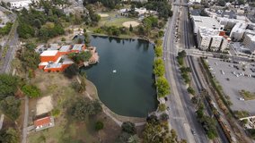 aerial footage of a silky green lake at Lincoln Park with cars and trucks driving along the street and office buildings in the skyline in Los Angeles California USA