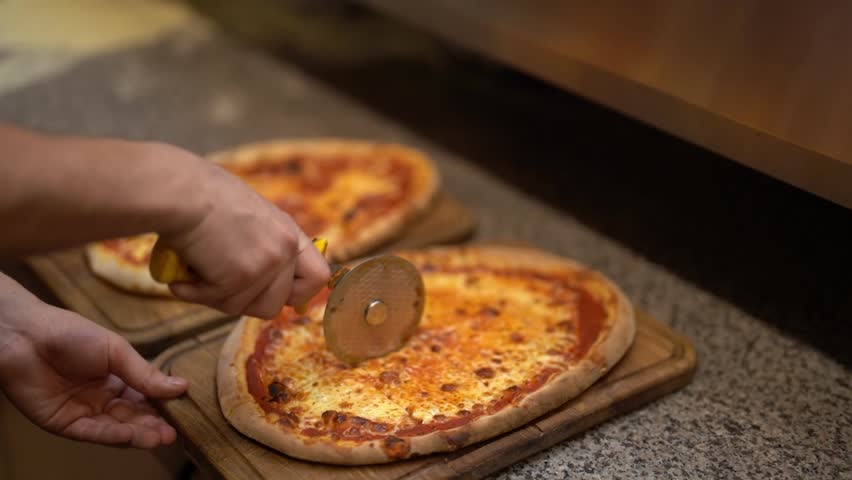 Side view from above of anonimous pizzeria worker slicing deliciously looking tomato cheese pizza with cutter wheel in order to be served