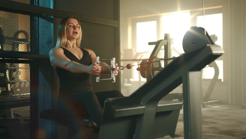 Pretty blonde with fit body in black sportswear performs seated cable row on cable machine. Woman exercises in modern fully-equipped gym lit by sunlight. High quality 4k footage