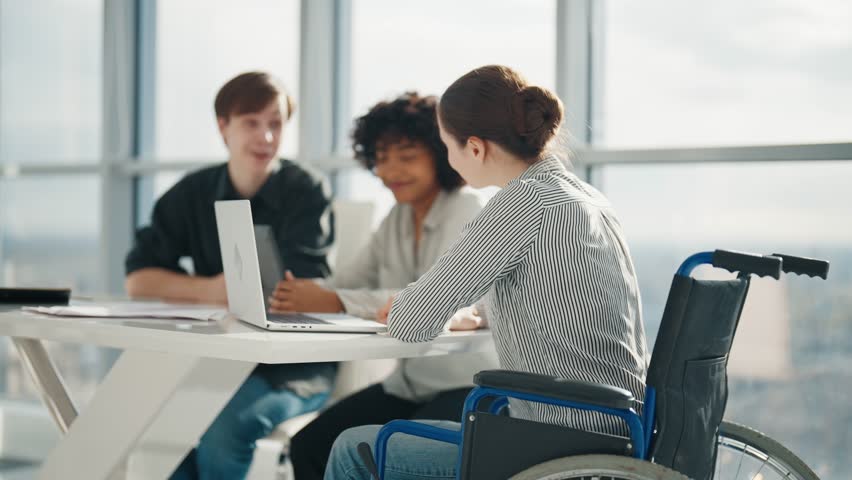 Friendly colleagues working on project in modern office together sitting at desk. Woman in wheelchair with physical disability on meeting with caucasian man african american female. Teamwork, discuss. Royalty-Free Stock Footage #1103943789
