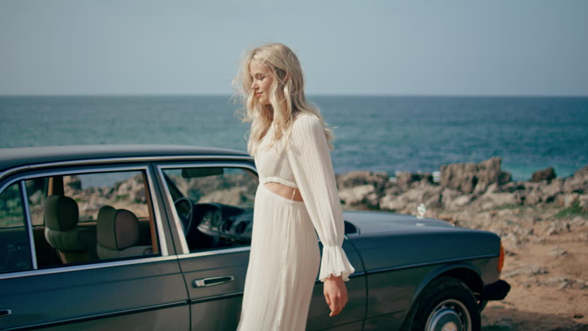 Woman posing leaning on car at beautiful ocean coast. Attractive carefree girl wearing white dress enjoy road trip on sunny seashore. Serene young blonde tourist standing near classic retro automobile Royalty-Free Stock Footage #1103946771