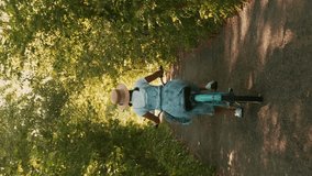 Female in straw hat and dress riding retro bicycle in forest. Vertical video. Brunette female moves on vintage bike along forest path, sunlights shining through tree leaves. Enjoying summer holidays