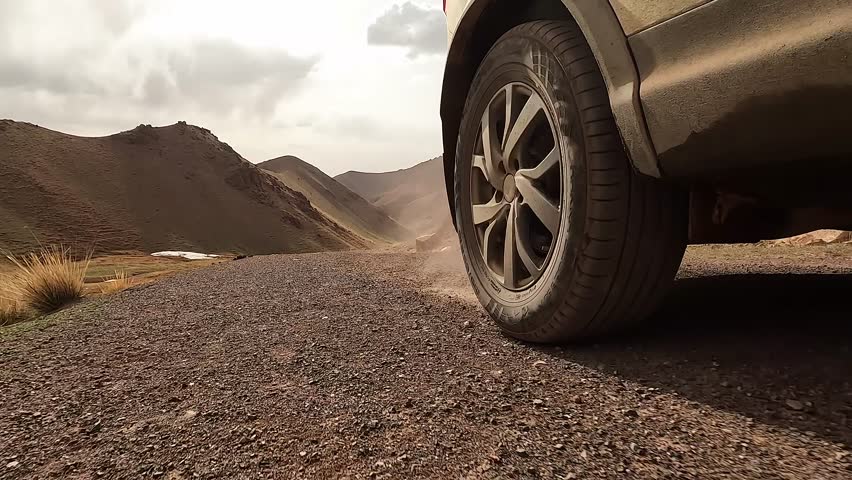Close-up of a car driving on a dirt rocky road, creating dust. View of the right rear wheel of an SUV in motion. Traveling by car in Kyrgyzstan. An exciting road adventure.