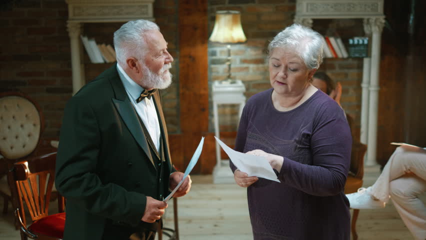 Medium shot of an old actor and elderly actress rehearsing a scene in the theater while holding their scripts