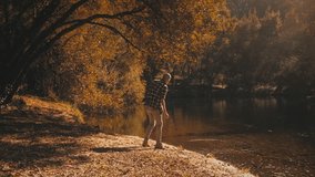 Man enjoys golden river and forest in autumn. He throws stones in water and listens to splash. Serene and soothing clip.