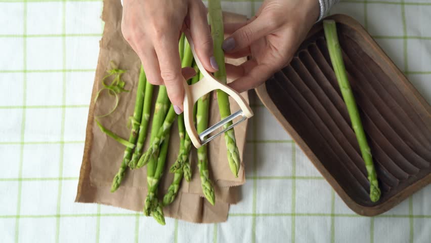 Girl hands peel the green asparagus - clean peel vegetable. Woman peeling the edible sprouts of asparagus with a vegetable peeler. Top view on spring raw ingredient and chef hand with knife. 4k 25FPS Royalty-Free Stock Footage #1103960301