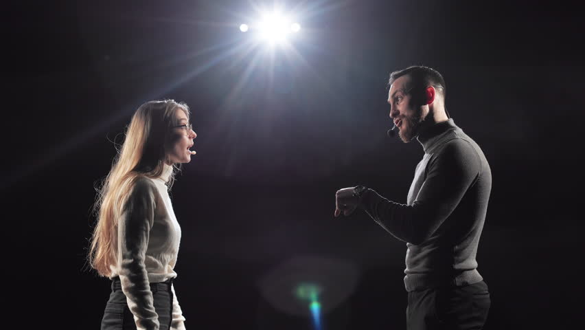 Side view of bearded man and beautiful woman having dispute during business performance on stage. Female leader in headset holding clipboard and making stop gesture with palm.