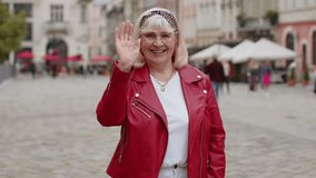 Senior woman smiling friendly at camera, waving hands gesturing hello, hi, greeting or goodbye, welcoming with hospitable expression outdoors. Elderly grandmother walking in urban city town street - Powered by Shutterstock - Get 15% off with code: PIKWIZARD15