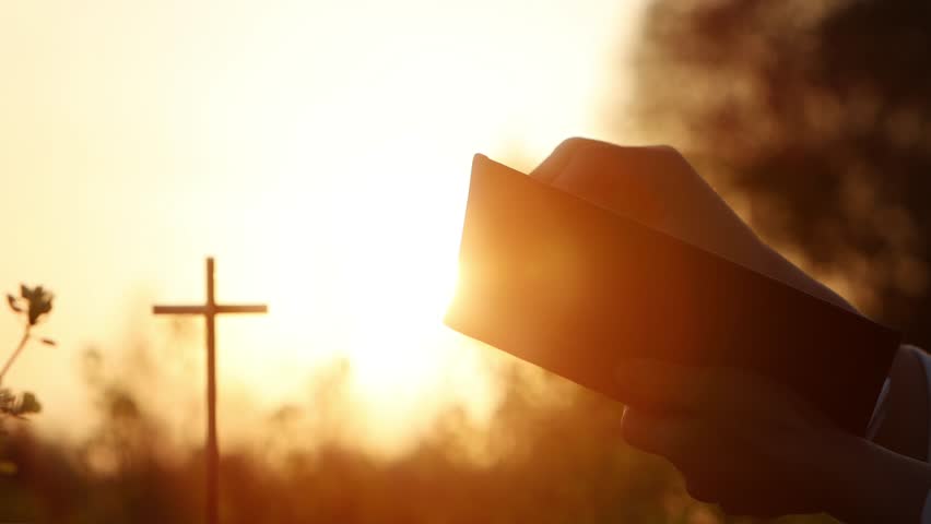 A Christian reading the Holy Bible in a beautiful evening with intense sunset light and a cross background symbolizing the death and resurrection of Jesus Christ
 Royalty-Free Stock Footage #1103976915