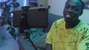 Medium close-up racking focus shot of smartphone on selfie stick, and young African American man sitting on couch and recording video content for his vlog on streaming platform