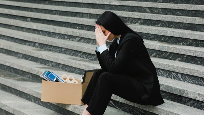 A brunette woman sits on the stairs with her head in her hands, feeling the weight of her unemployment and loss of work. Unhappy Professional Sitting on Stairs After Job Loss Royalty-Free Stock Footage #1103979639