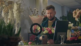 Male florist collects bouquet of flowers in flower store, shoots video for blog using ring lamp, smartphone and digital tablet. Concept of floristry, retail floral small business and entrepreneurship.