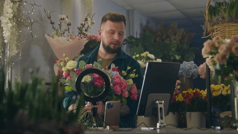 Male florist collects bouquet of flowers in flower shop, records video for blog using ring lamp, smartphone and digital tablet. Concept of floristry, retail floral small business and entrepreneurship. Arkivvideo