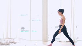 Zoom out attractive sporty Asian woman practicing yoga, doing Virabhadrasana 1 exercise, meditating in Warrior One yoga pose, indoor working out at home, wearing sportswear. Full length video.
