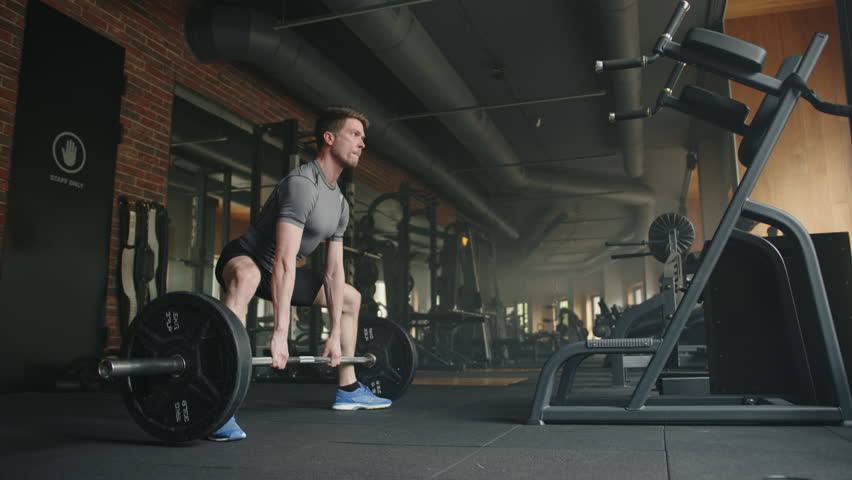 Young handsome athletic man lifts barbell in spacious gym with strength training equipment in black colors. Sportsman performs deadlift, legs far apart. High quality 4k footage Royalty-Free Stock Footage #1103991079