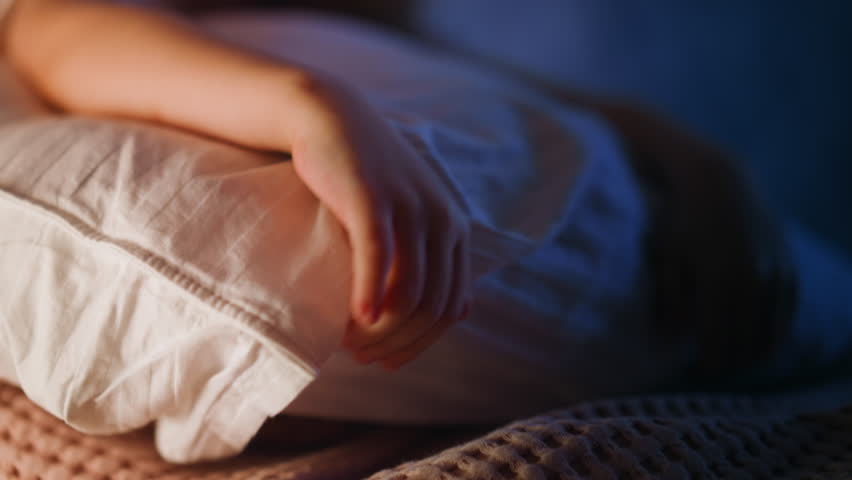 Unhappy little girl with large white pillow wipes away tears sitting alone on bed at lamp and moon lights in empty bedroom closeup slow motion Royalty-Free Stock Footage #1103993901