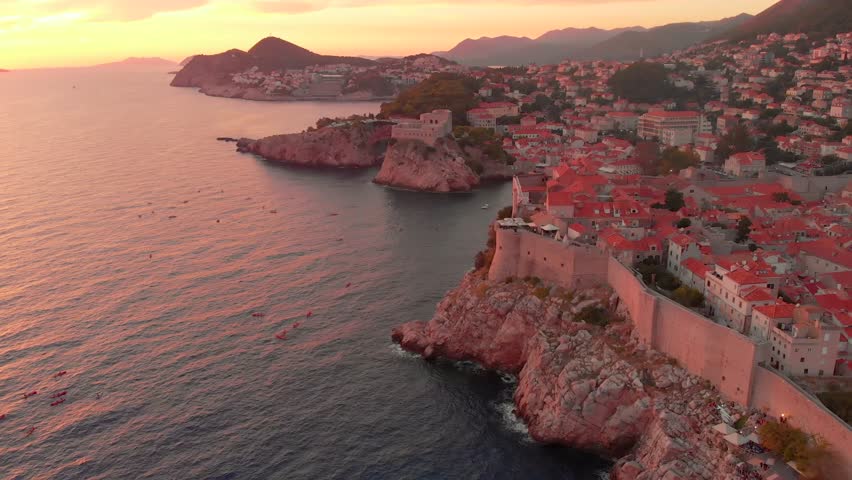 An aerial view of a sunset over Dubrovnik. People can be seen enjoying the sunset on kayaks in front of the city walls. The walls of the old town visible