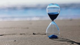 Hourglass with blue sand inside in a warm golden morning sunlight with shadow on a sandy beach and sea background, starting time for a new day or running of time. Concept of the rapid passage of time. - Powered by Shutterstock - Get 15% off with code: PIKWIZARD15