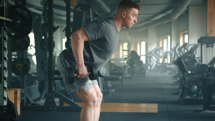 Profile shot of young athlete performing bent over dumbbell row in gym. Sportsman with slightly bent knees leans forward and pulls the weight up to waist. High quality 4k footage