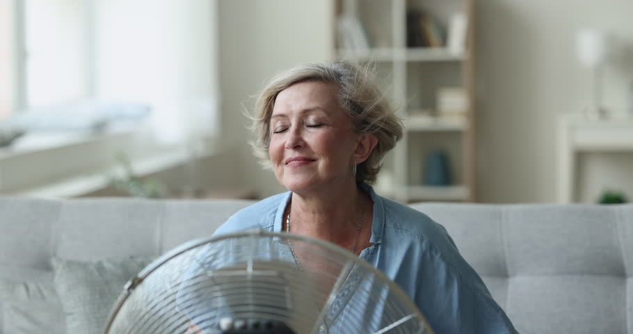 Happy relaxed senior lady sitting at fresh cool air blowing from fan, resting on home sofa, enjoying freshness from electric blower, ventilator, cooler, using home appliance, equipment for ventilation Royalty-Free Stock Footage #1104029177
